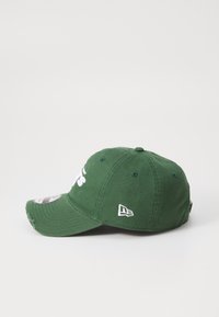 Grønn caps med hvit brodert tekst, en buet visir og en logo på siden. Har en teksturert stoff og synlige sømdetaljer.