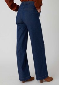 Pantaloni in denim scuro a gamba larga con una texture morbida, dotati di tasche posteriori e dettagli di cucitura discreti, che si abbinano a stivali alla caviglia in suede marrone.