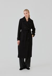 Black wool trench coat with double-breasted buttons, a belt, wide collar, and long sleeves. Paired with black pants and shoes.