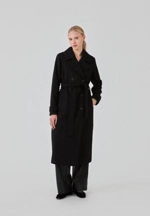 Svart ull trenchcoat med dubbelknäppning, bälte, bred krage och långa ärmar. Matchas med svarta byxor och skor.