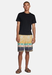 Maglietta nera, pantaloni da bagno a motivi con sole giallo e palme in blu, verde e rosa. Infradito marroni completano il look.