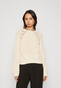 Cremefarvet strikket sweater med rund halsudskæring, tekstureret krop, blonde detaljer ved ærmerne og ribbede manchetter, kombineret med sorte bukser.