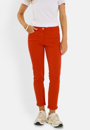 Femme portant un pantalon orange vif retroussé, une chemise blanche à manches courtes, des baskets beiges et des bracelets dorés, debout avec une main dans la poche.
