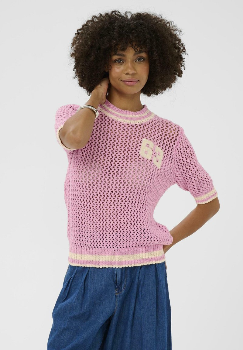 Kõrgete lokkis juustega naine, kes kannab roosat lühikeste varrukatega kootud tualetti numbrimärgiga "63" ja siniseid laiade jalgadega pükse, poseerides käega kaela juures.