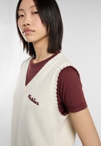 Chaleco de punto crema con cuello en V y bordes acanalados, superpuesto a una camiseta burdeos. Logo "Dickies" bordado en la parte frontal del chaleco.