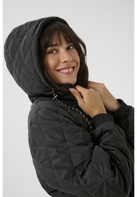 Schwarze gesteppte Jacke mit Kapuze, aus einem strukturierten Stoff, mit weißen Zugbändern und gerippten Bündchen. Das Design ist lässig und figurbetont.