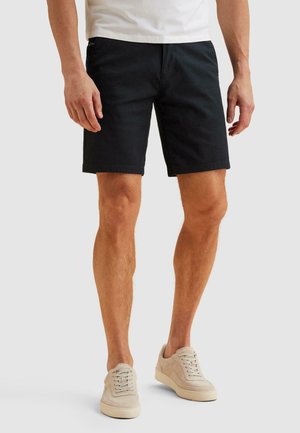 Mann trägt schwarze knielange Shorts, weißes T-Shirt und beige Turnschuhe und steht vor einem einfarbigen Hintergrund.