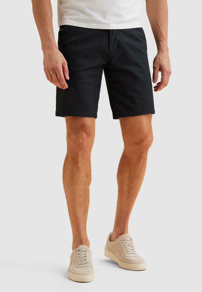 Homme portant un short noir au genou, un t-shirt blanc et des baskets beiges, debout devant un fond uni.