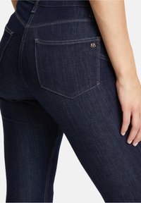 Jeans in denim blu scuro con una vestibilità attillata, dotati di una tasca posteriore sagomata e un piccolo logo decorativo. La texture appare liscia e leggermente elastica.