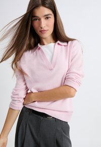 Jasnoróżowy sweter dzianinowy z dekoltem w serek i podwiniętymi mankietami, zestawiony z szarymi, dopasowanymi spodniami. Gładka tekstura, swobodny fason.