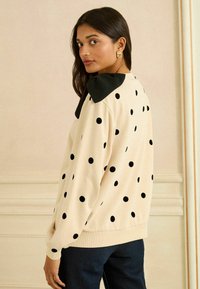 Krémový sveter s veľkými čiernymi bodkami, s kontrastným čiernym golierom. Mäkká tkanina a voľný strih, s rebrovanými manžetami a lemom.