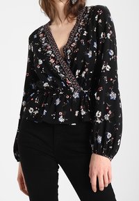 Blusa avvolgente nera a fiori con maniche lunghe a sbuffo, scollatura a V e vita elastica. Presenta motivi floreali blu, rossi e bianchi e un bordo decorativo.