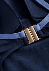 Primer plano de tela azul marino con correas azul claro y una hebilla rectangular de color dorado que asegura la prenda.