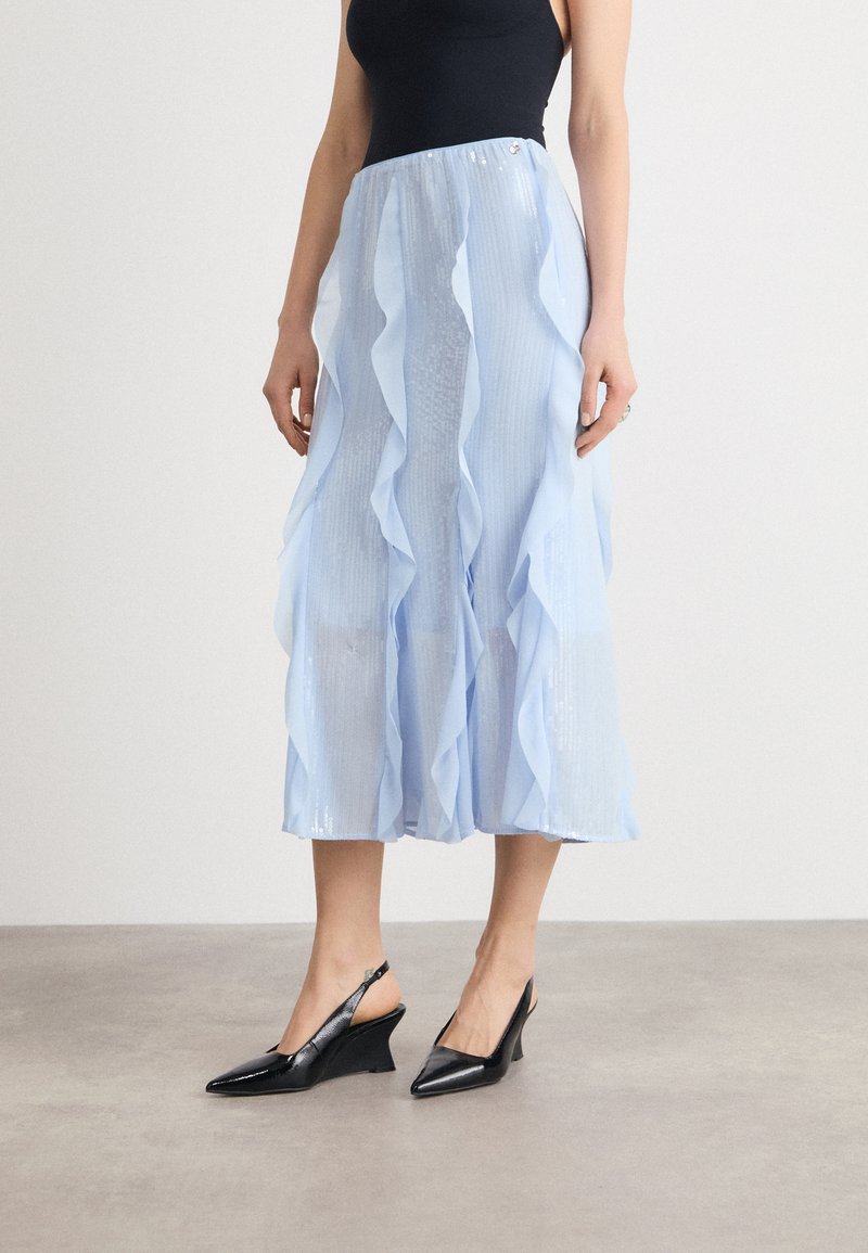 Jupe midi plissée bleu clair avec des couches froncées et un éclat subtil, associée à des talons compensés à brides noires avec des bouts pointus.