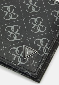 Portefeuille noir en simili cuir avec un motif logo gris, des accents de couture diagonale, et un détail en métal argenté en forme de triangle "Guess ?".