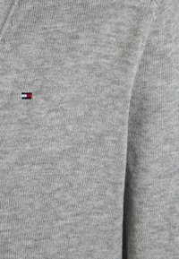 Hellgrauer, gestrickter Pullover mit V-Ausschnitt und kleinem, gesticktem Logo in Rot, Weiß und Blau auf der linken Brustseite.
