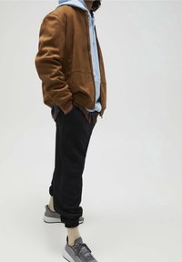 Bruin suède bomberjack over een lichtblauwe hoodie, gecombineerd met zwarte joggers en grijze sneakers. Neutrale kleuren, casual pasvorm, gestructureerde materialen.