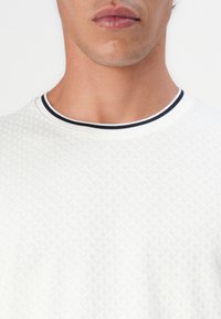 T-shirt bianco con texture e un design a maglia motivo. Presenta un collo rotondo e un colletto a coste bianco e nero a contrasto.