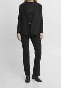 Osoba w czarnym żakiecie, czarnym topie, czarnych dopasowanych spodniach, czarnych botkach na obcasie i srebrnej bransolecie na lewej ręce.