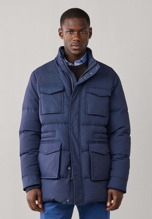Joven con una chaqueta de invierno acolchada azul con cuatro bolsillos delanteros con solapa y un cuello alto, de pie frente a un fondo liso.