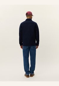 Navy blue jacket with long sleeves and button closure, paired with blue denim jeans. The person wears a maroon cap and brown shoes.