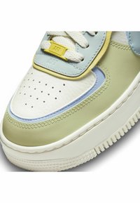 Närbild av en pastellfärgad sneakers tå med vita snören, gul snörtag, perforerat vitt läder, gröna och blå paneler, samt strukturerad sula.