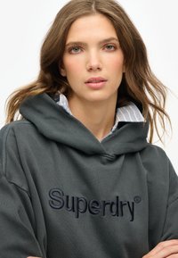 Felpa con cappuccio grigio scuro con logo "Superdry" ricamato in blu navy. Presenta un grande cappuccio e un colletto a strati con righe blu e bianche.