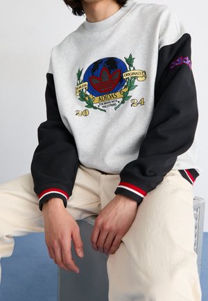 Personne portant un sweat Adidas avec un globe, un logo trèfle rouge, le texte "20 24" et "Adidas Originals", assise les mains reposant sur les genoux.