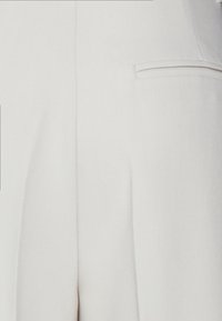 Pantalones de color gris claro con pliegues, confeccionados en una tela suave. Cuentan con un bolsillo trasero con un borde sutil. Diseño sencillo y ajustado.