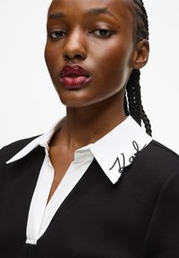 Čierny odev s bielym golierom, na ktorom je vyšívané logo "Karl". Hladká textura, design s výstrihom do V a viditeľný detail vrkoča.