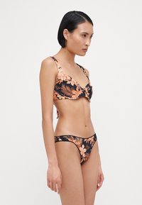 Fekete virágmintás bikini barack és bézs trópusi mintákkal. A melltartón hátkötő található; az alsó rész alacsony derékkal és minimális fedéssel rendelkezik.