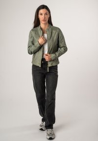 Giacca di pelle verde oliva con collo alto, chiusura con zip e due tasche frontali, abbinata a jeans neri a gamba larga e sneakers.
