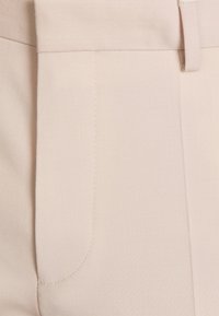 Gros plan sur un pantalon beige clair montrant la poche avant, les coutures et les détails de la ceinture sur un tissu lisse.