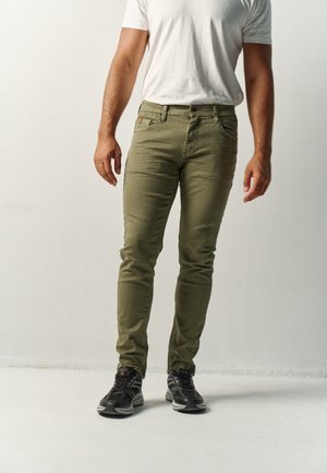 Hombre con vaqueros ajustados verde oliva, camiseta blanca y zapatillas negras, de pie frente a un fondo claro y liso.
