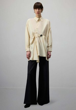 Kobieta w oversize kremowej koszuli zapinanej na guziki z marszczonym detalem i czarnych szerokich spodniach, stojąca na jednolitym szarym tle.