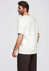 Muž s vousy a tetováním na levém ruce, oblečený v jednoduché bílé košili s krátkým rukávem a tmavých kalhotách, stojící na jednobarevném pozadí.