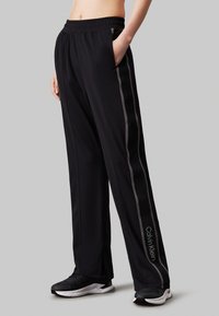 Pantaloni sportivi neri con vestibilità rilassata, dotati di vita con cordino, tasche laterali e righe grigie. Logo visibile sulla parte bassa della gamba.