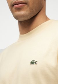 Κοντινό πλάνο στο κάτω μέρος του προσώπου και τον λαιμό ενός ατόμου που φοράει μια μπεζ μπλούζα με ένα μικρό πράσινο κεντημένο λογότυπο κροκόδειλου στο στήθος.