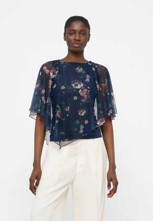 Kobieta ubrana w przezroczystą granatową bluzkę w kwiaty z falbanowymi rękawami i białe plisowane spodnie z wysokim stanem, stojąca na jednolitym tle.