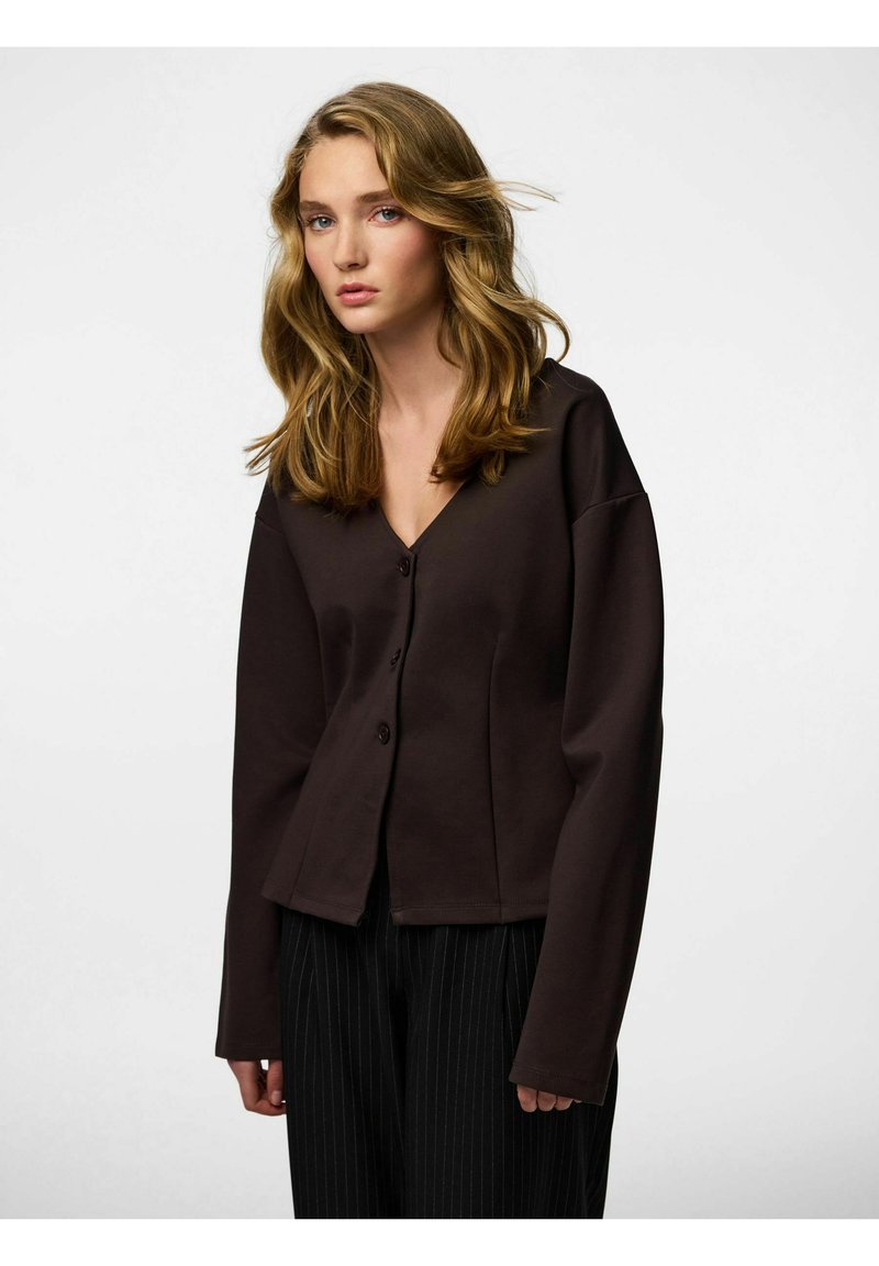 Hnědý přiléhavý cardigan s výstřihem do V, dlouhými rukávy a třemi knoflíky vpředu. Vyrobený z hladké látky, v kombinaci s kalhotami s tenkými pruhy.