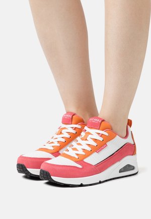 Des pieds portant des sneakers Skechers rose, orange et blanc avec des lacets blancs et une semelle à coussin d'air visible sur un fond blanc.