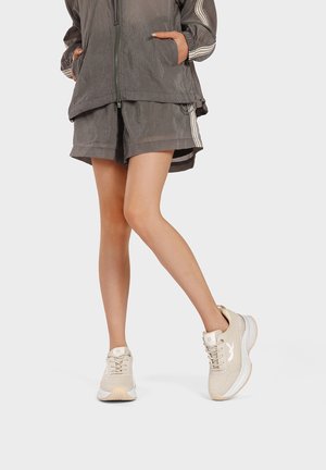 Persona con pantaloncini grigi e giacca con zip abbinata, scarpe da ginnastica beige con lacci e suole bianche, in piedi con una gamba incrociata sull'altra.