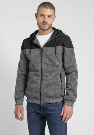 Homme portant un sweat à capuche gris zippé avec une partie supérieure noire et un jean bleu, debout devant un fond clair uni.