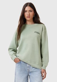 Felpa verde menta con una vestibilità rilassata, collo a girocordo a coste e logo ricamato. Indossata con jeans di denim blu chiaro. Tessuto morbido.