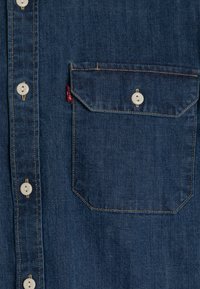 Donkerblauwe denimshirt met witte knopen, een borstzak en zichtbare stikseldetails. Bevat een merketiket op de zak.