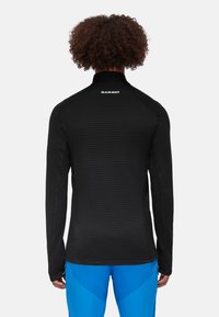 Black long-sleeve shirt with a textured, ribbed design and high collar. Logo on the upper back, paired with blue athletic pants.