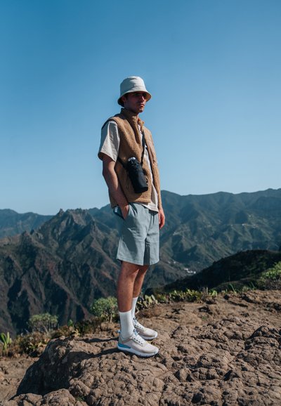Light brown fleece vest, white t-shirt, light blue shorts, white socks, and gray sneakers with blue accents, set against a mountainous background.
