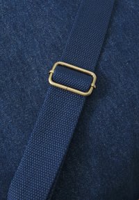 Navy strapmateriaal met een gestructureerde afwerking, met een rechthoekige messing gesp die de strap vastmaakt aan een denim-achtig stoffen achtergrond.