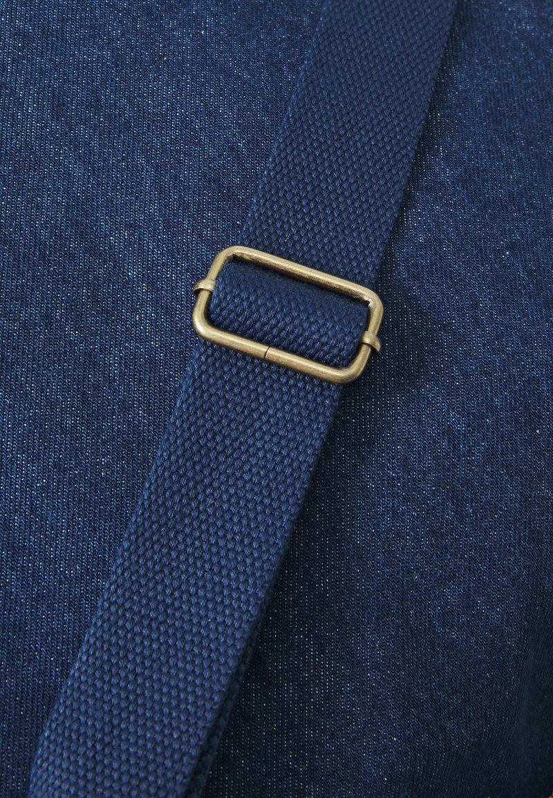 Navy strapmateriaal met een gestructureerde afwerking, met een rechthoekige messing gesp die de strap vastmaakt aan een denim-achtig stoffen achtergrond.