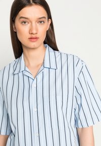 Camisa de meia manga azul clara com riscas verticais em azul marinho, frente com botões, gola e um único bolso no peito. Textura de tecido suave.